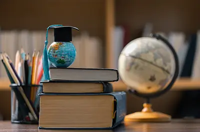 A small globe with a graduation cap rests on a stack of books on a desk. A pencil holder filled with colored pencils is nearby, and a larger globe is slightly blurred in the background. Shelves with books are visible behind.