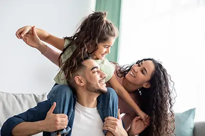A joyful family moment: a man gives a young girl a piggyback ride while a woman smiles warmly at them. The girl, with her hair in a ponytail, happily interacts with the man. They are in a bright room with a light-colored sofa.