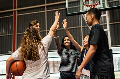 A group of four young people standing on a basketball court, giving each other high-fives. One person holds a basketball. A hoop and net are visible in the background. They are smiling and appear to be in a celebratory mood.