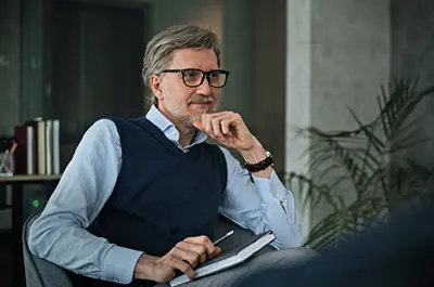 A man with gray hair and glasses sits thoughtfully in an office chair, holding a notebook and pen. He is wearing a blue sweater over a button-down shirt. In the background, there are books and a potted plant.
