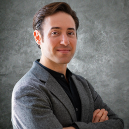 A man with short brown hair wearing a grey blazer and black shirt stands confidently with arms crossed, smiling slightly, in front of a textured gray background.